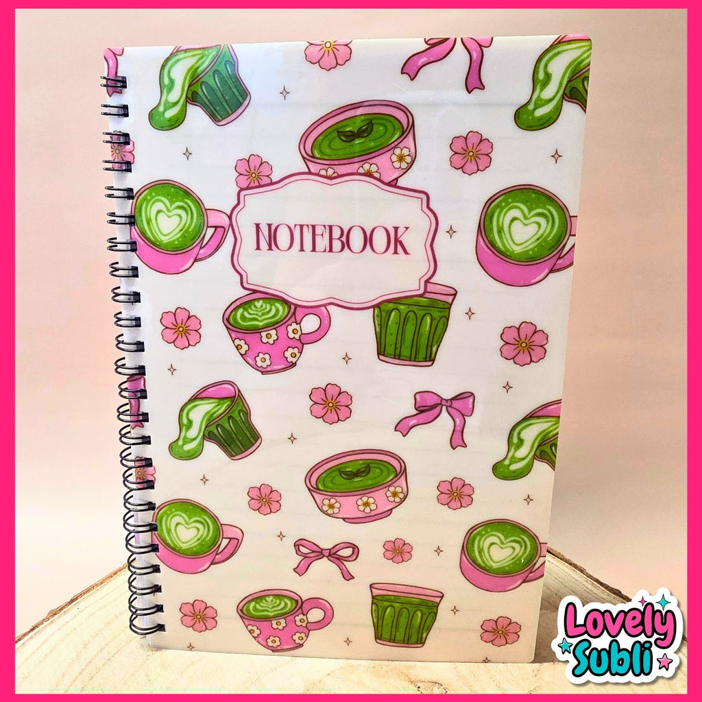 A spiral-bound notebook with a white cover decorated with pink and green illustrations of coffee cups, cupcakes, bows, and flowers, featuring a central label that reads 'NOTEBOOK'.