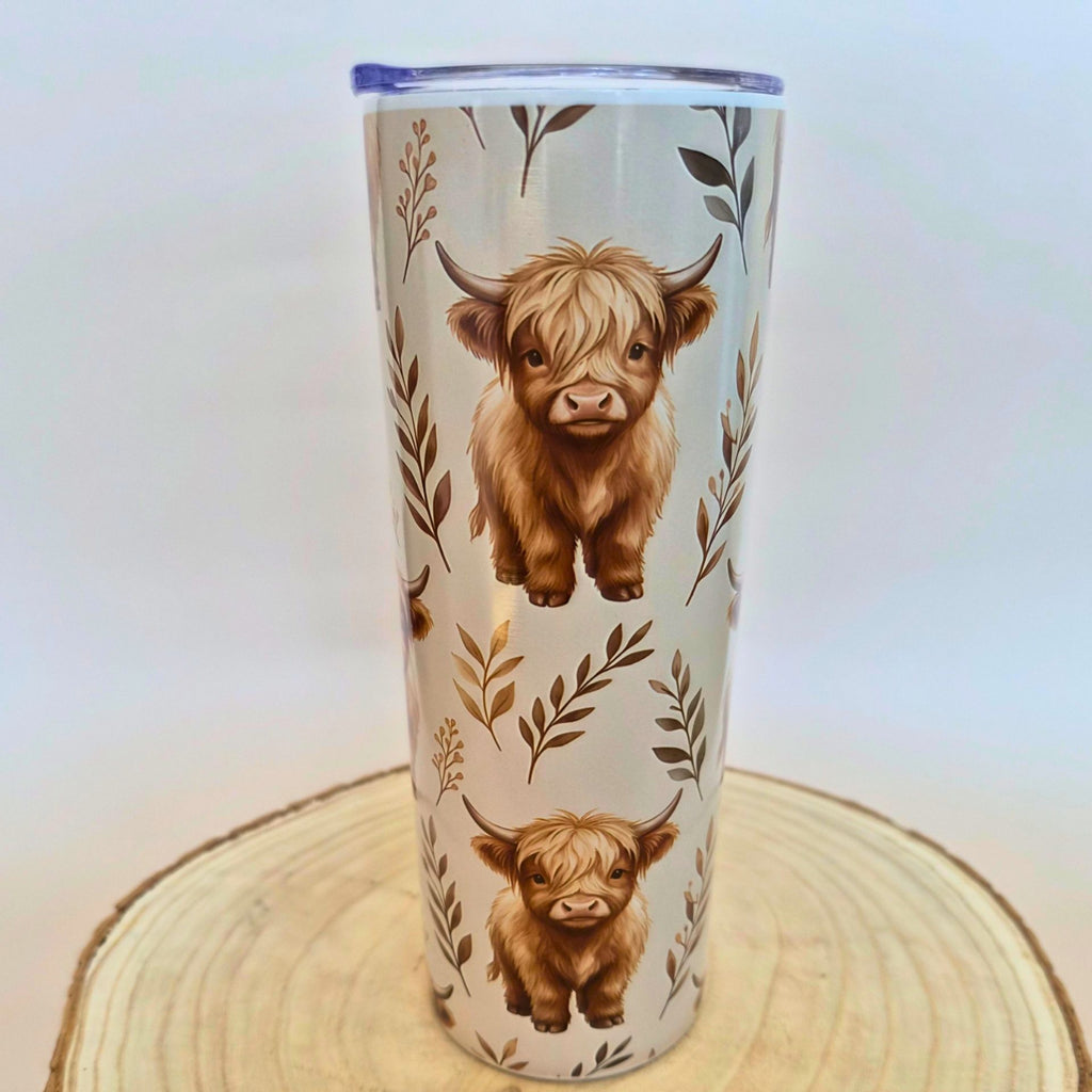 A tall insulated tumbler with a clear lid, decorated with two brown Highland cow illustrations and botanical leaf motifs on a light background, displayed on a round wood slice coaster.