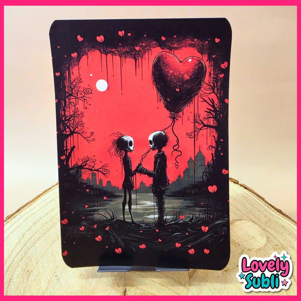 A vertical art print or card featuring a gothic illustration of two skeletal figures holding hands under a large heart balloon, set against a red sky with black trees and scattered red hearts. The print is displayed upright on a small stand on a wooden surface, with a pink border and a 'Lovely Subli' logo in the bottom right corner.
