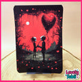 A vertical art print or card featuring a gothic illustration of two skeletal figures holding hands under a large heart balloon, set against a red sky with black trees and scattered red hearts. The print is displayed upright on a small stand on a wooden surface, with a pink border and a 'Lovely Subli' logo in the bottom right corner.