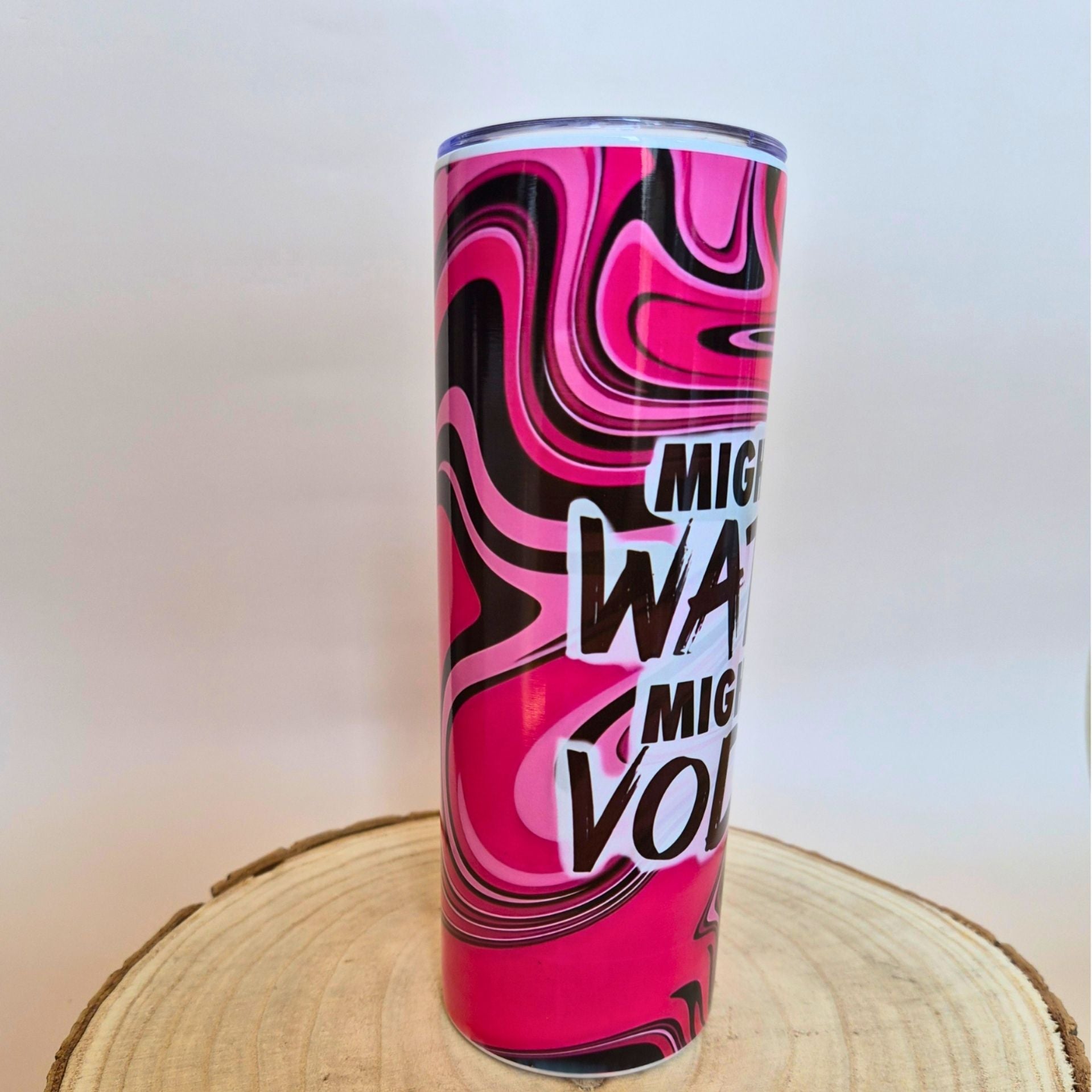A tall insulated tumbler with a vibrant pink, black, and white marbled swirl pattern and bold text reading 'Mighty Water Volume' in white and black, displayed on a round wooden slice against a plain light background.