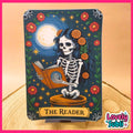 A decorative art print or card titled "THE READER" featuring a Day of the Dead style skeleton seated on a stool and reading a book, with a large moon, flowers, stars, and green vines on a dark blue background. The piece is displayed upright on a small stand on a wood surface against a warm beige backdrop, with a pink border and a "Lovely Subli" logo in the lower right.