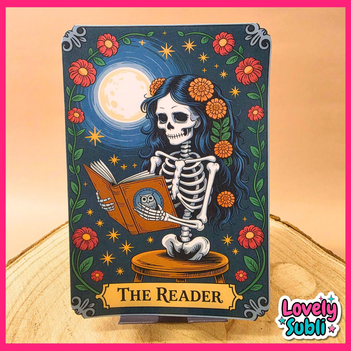 A decorative art print or card titled "THE READER" featuring a Day of the Dead style skeleton seated on a stool and reading a book, with a large moon, flowers, stars, and green vines on a dark blue background. The piece is displayed upright on a small stand on a wood surface against a warm beige backdrop, with a pink border and a "Lovely Subli" logo in the lower right.
