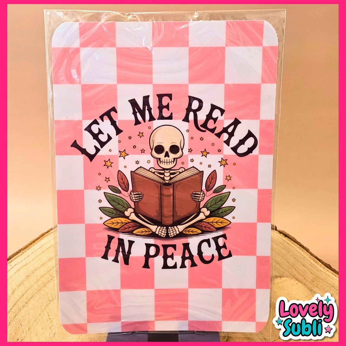 A square printed sign or sticker featuring a pink and white checkered background with an illustration of a skeleton reading a book, surrounded by the words “LET ME READ IN PEACE.” The item is photographed inside a clear plastic sleeve, standing on a small black base against a warm beige background.