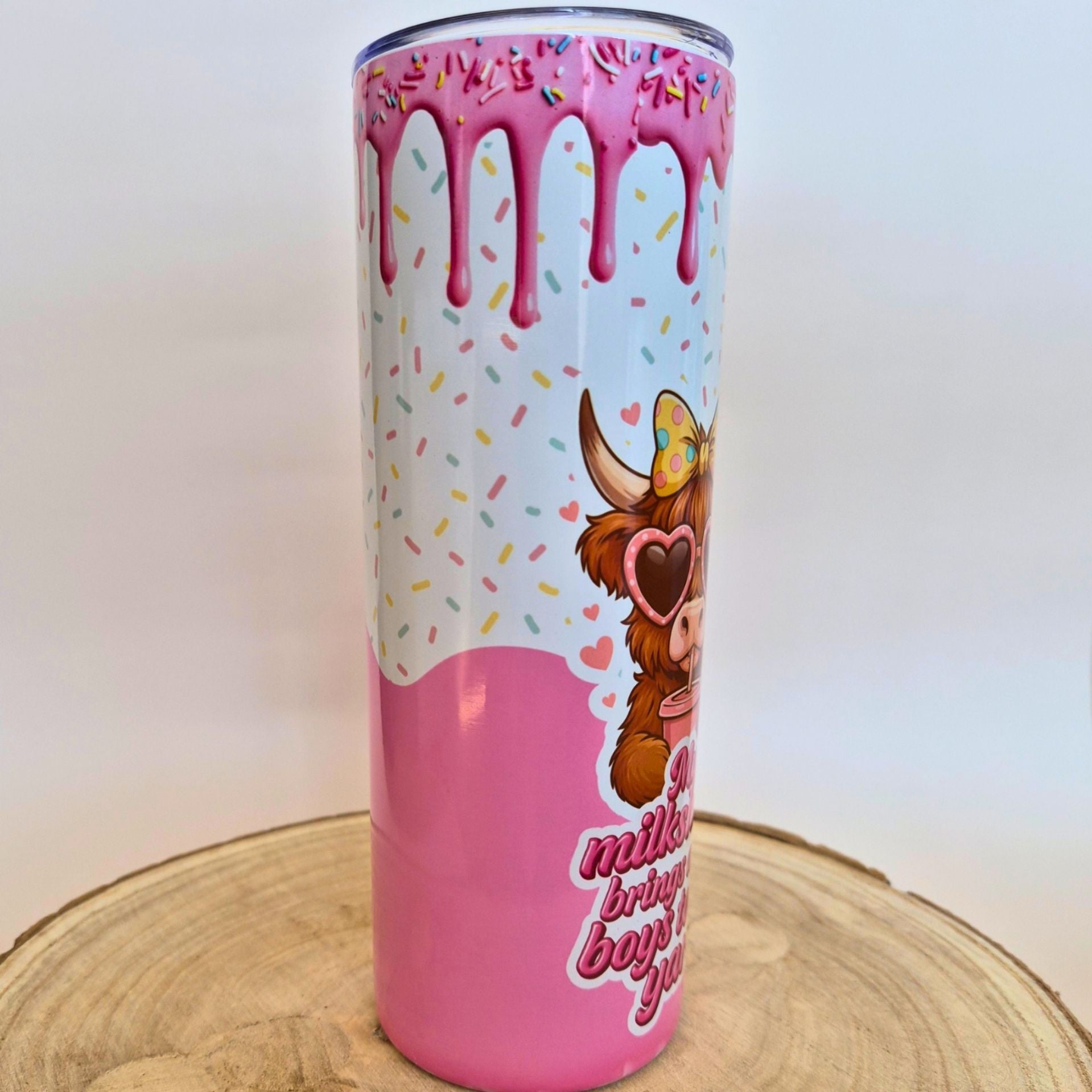 A tall insulated tumbler with a pink and white donut-themed design, featuring dripping pink glaze, colorful sprinkles, a cartoon cow wearing heart-shaped sunglasses, and partial text on a light wood slice background.