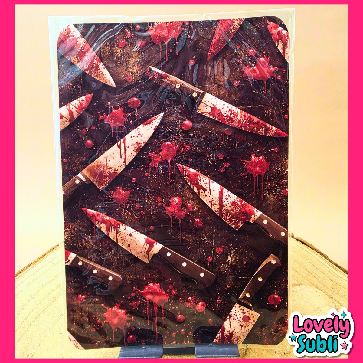 A rectangular art print or card featuring a dark, bloody scene with multiple knives and red splatter, displayed on a small stand against a light wood surface. The print has a white border and a 'Lovely Subli' logo in the bottom right corner.