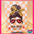 A rectangular printed card or sign with a true crime themed illustration, standing on a small black display stand. The design shows a woman's face with dark hair in a bun, yellow crime scene tape, red blood splatter details, and the text 'TRUE CRIME obsessed' on an orange-and-white checkered background.