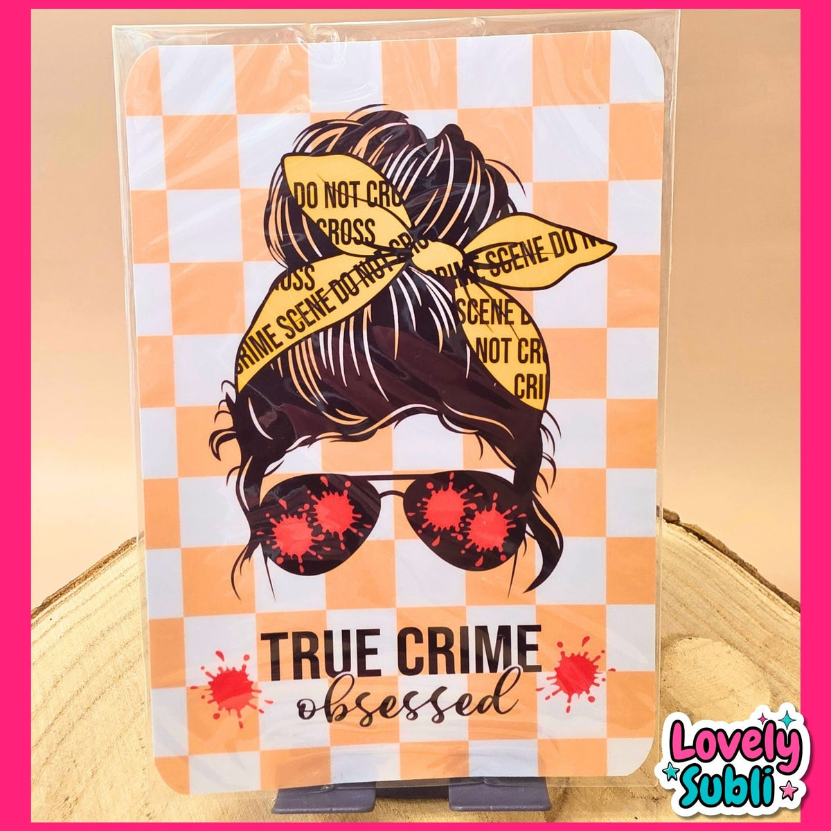 A rectangular printed card or sign with a true crime themed illustration, standing on a small black display stand. The design shows a woman's face with dark hair in a bun, yellow crime scene tape, red blood splatter details, and the text 'TRUE CRIME obsessed' on an orange-and-white checkered background.