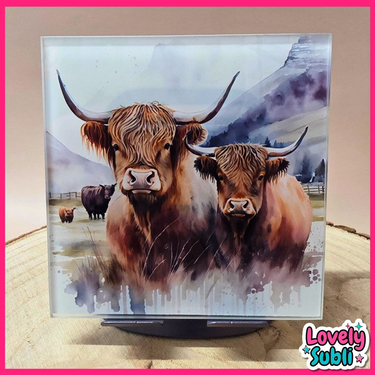A square acrylic art plaque featuring a watercolor painting of two Highland cows in a misty mountain landscape, displayed on a stand against a neutral background with a pink border.
