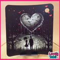 A square black decorative plaque or coaster featuring a dark fantasy illustration of a large dripping heart balloon above a silhouetted couple in a graveyard, with small red hearts floating in the sky. The item is displayed on a small stand on a wooden surface against a beige background.