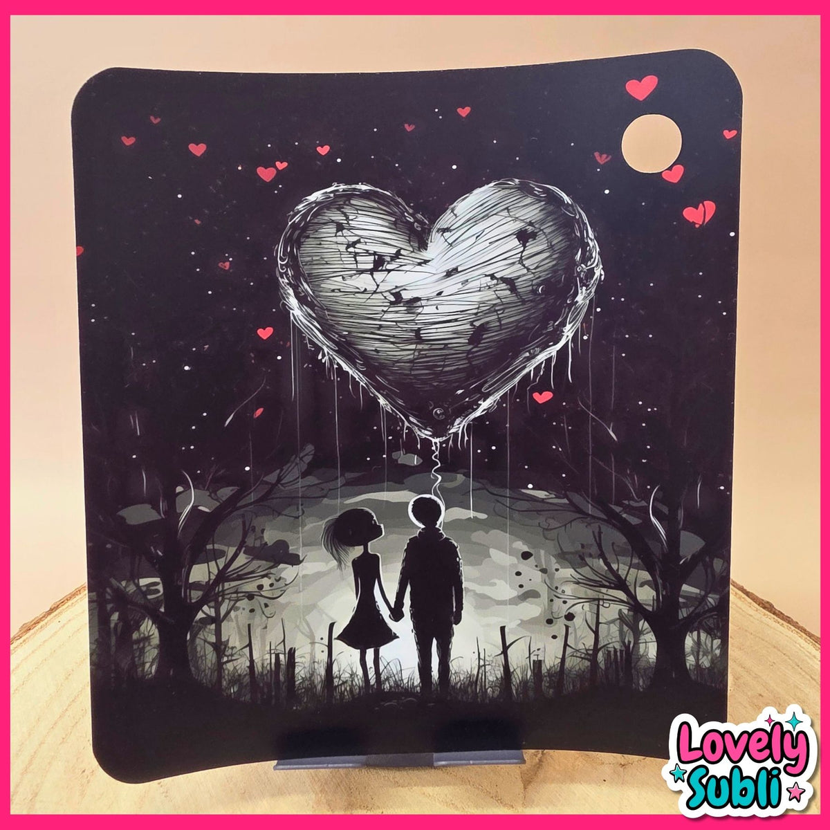 A square black decorative plaque or coaster featuring a dark fantasy illustration of a large dripping heart balloon above a silhouetted couple in a graveyard, with small red hearts floating in the sky. The item is displayed on a small stand on a wooden surface against a beige background.