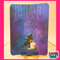 A rectangular decorative board or plaque with rounded corners, featuring a dark blue and purple celestial design with a raven holding a candle, a pentagram symbol, stars, and a crescent moon. The board is displayed on a small stand on a wooden surface, framed by a bright pink border.