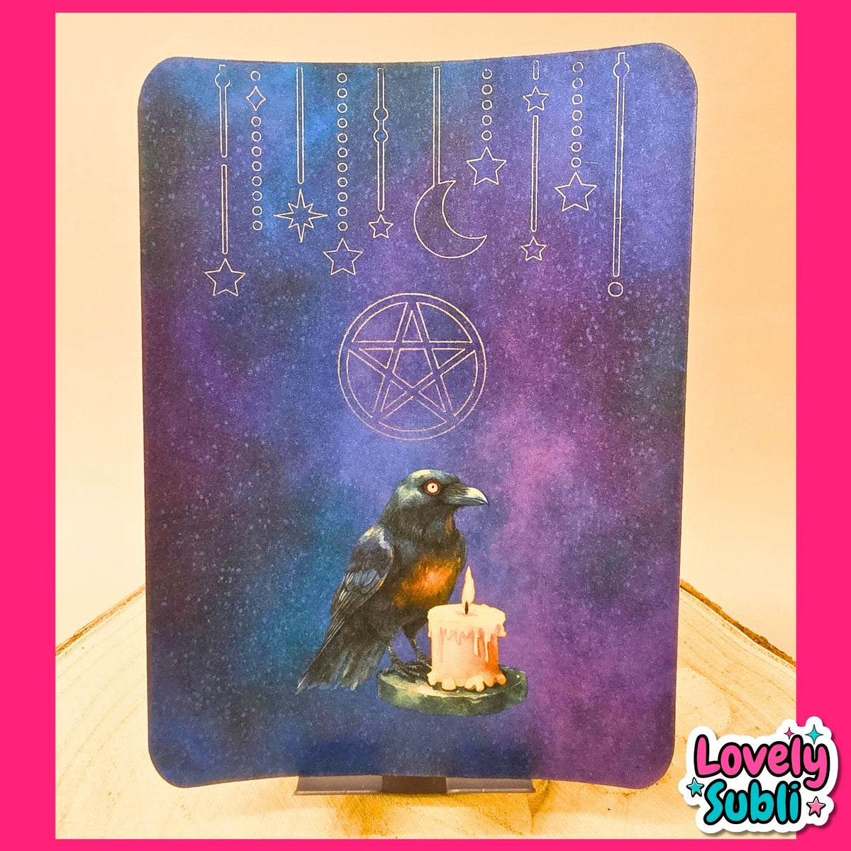 A rectangular decorative board or plaque with rounded corners, featuring a dark blue and purple celestial design with a raven holding a candle, a pentagram symbol, stars, and a crescent moon. The board is displayed on a small stand on a wooden surface, framed by a bright pink border.