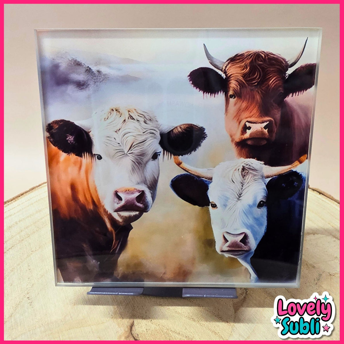 A square acrylic art plaque featuring a painted portrait of three cows, displayed on a small stand. The artwork has a white border and is photographed on a light wood surface with a pink background.