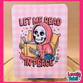 A rectangular art print or card with rounded corners featuring a pink gingham border and a cartoon skeleton in a pink hoodie reading a book, with the text 'LET ME READ IN PEACE.' The print is photographed standing upright on a small stand against a warm tan background, with a bright pink outer border and a 'Lovely Subli' logo in the lower right.