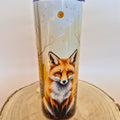 A tall insulated tumbler with a lid, featuring a detailed illustration of a red fox in a light-colored forest scene, displayed on a round wooden slice against a plain light background.