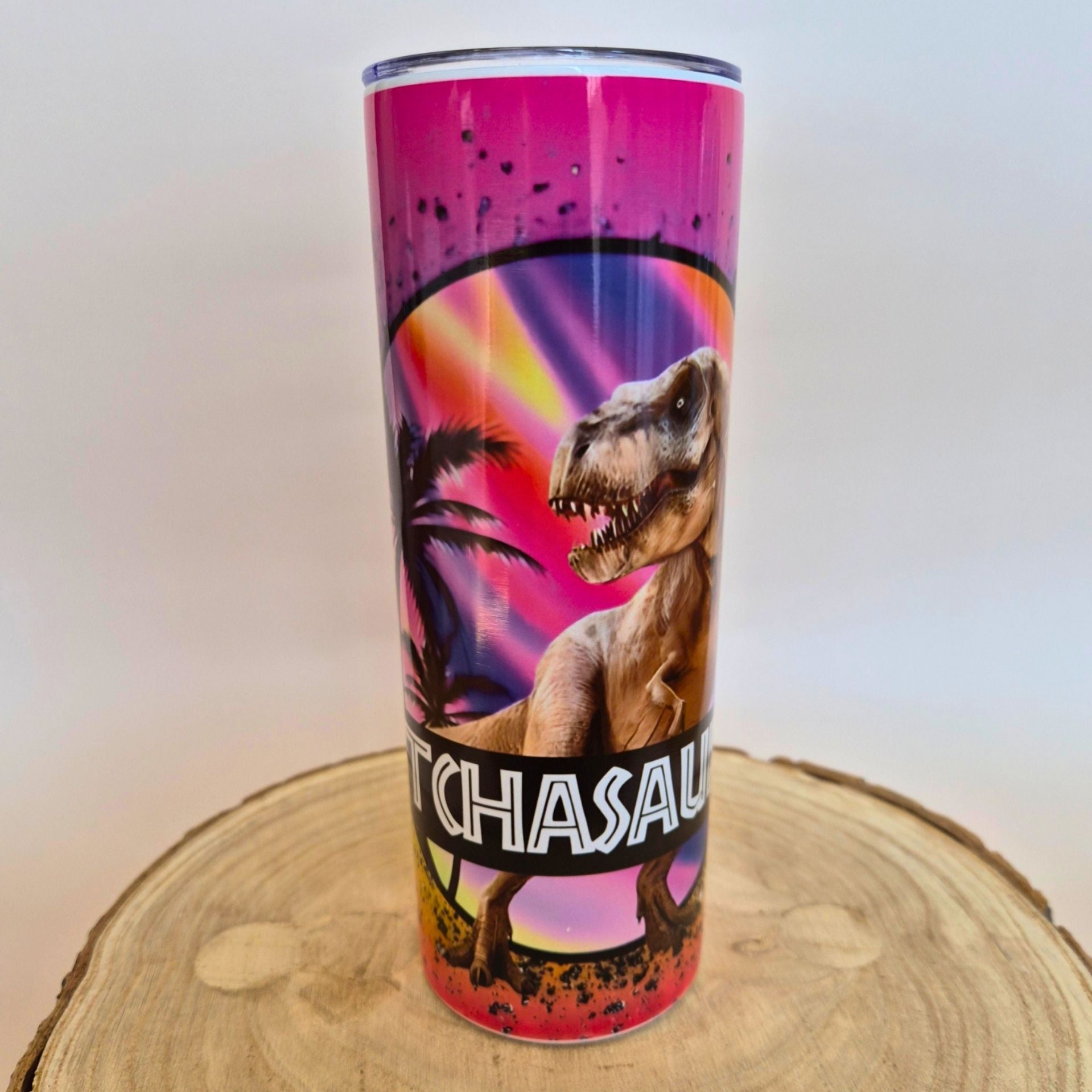 A tall insulated tumbler with a clear lid, featuring a colorful dinosaur graphic and the text 'TCHASAU' on the front, displayed on a round wood slice against a plain light background.