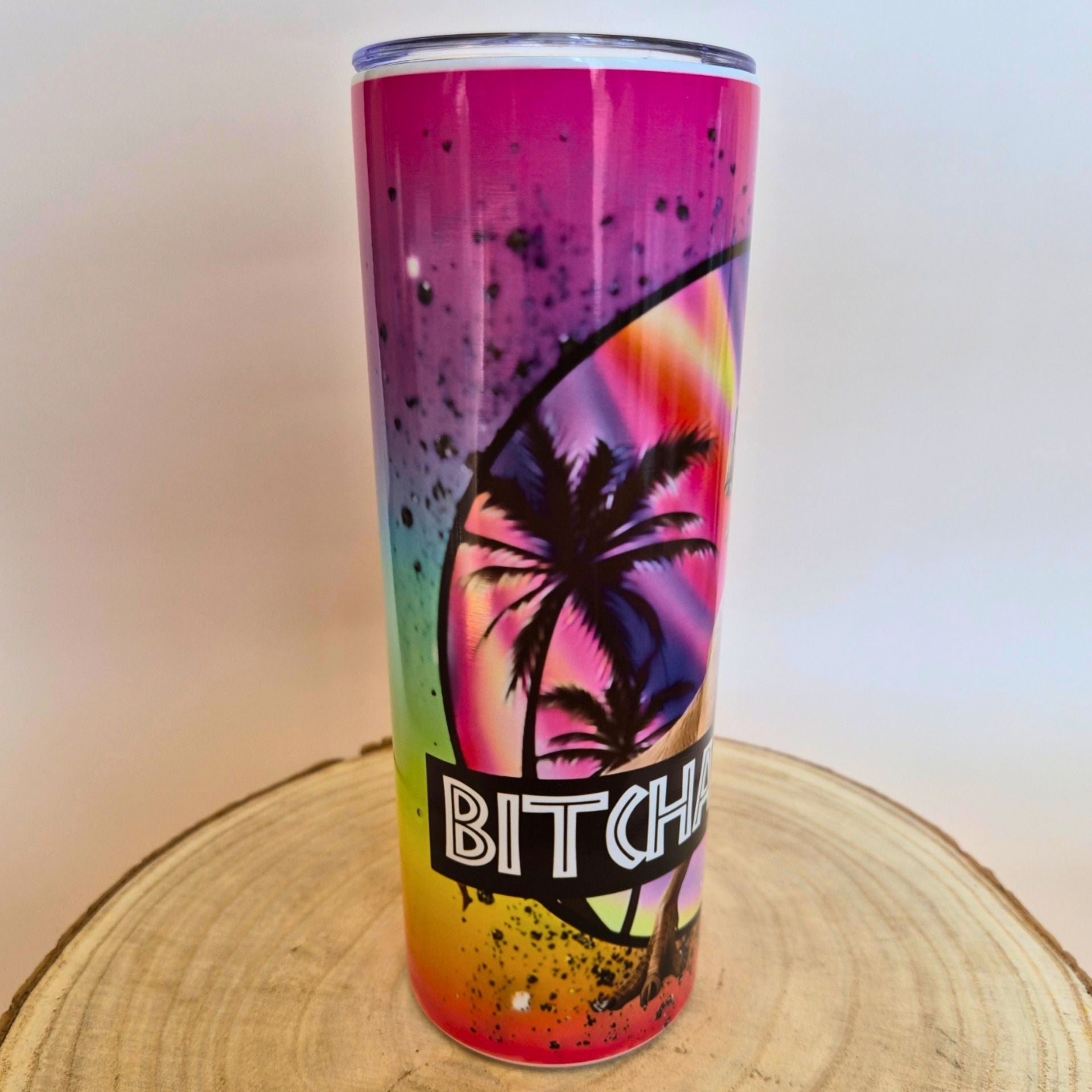 A tall insulated tumbler with a vibrant rainbow gradient and black palm tree graphic, sitting on a round wooden slice against a plain light background.