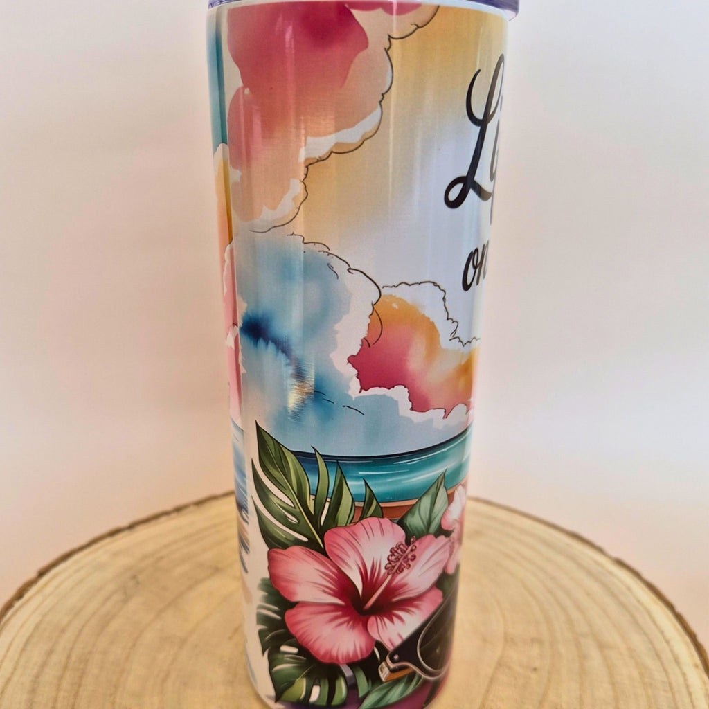 A tall insulated tumbler with a tropical beach and floral design, featuring pink hibiscus flowers, palm leaves, a blue ocean, and a sunset sky. The tumbler is shown standing on a circular wooden slice against a plain light background.