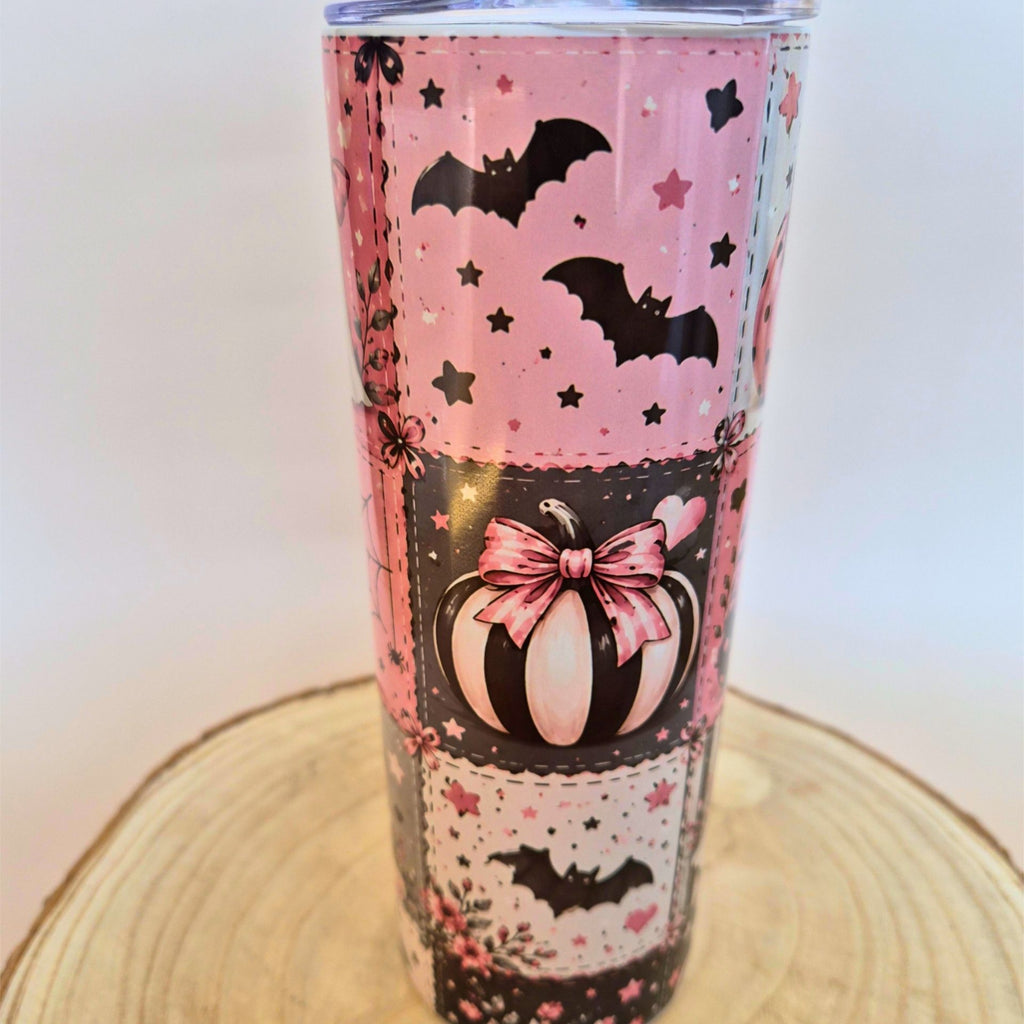 A pink and black Halloween-themed tumbler with bats, stars, a pumpkin, and floral lace patterns, shown on a wooden slice coaster against a light background.