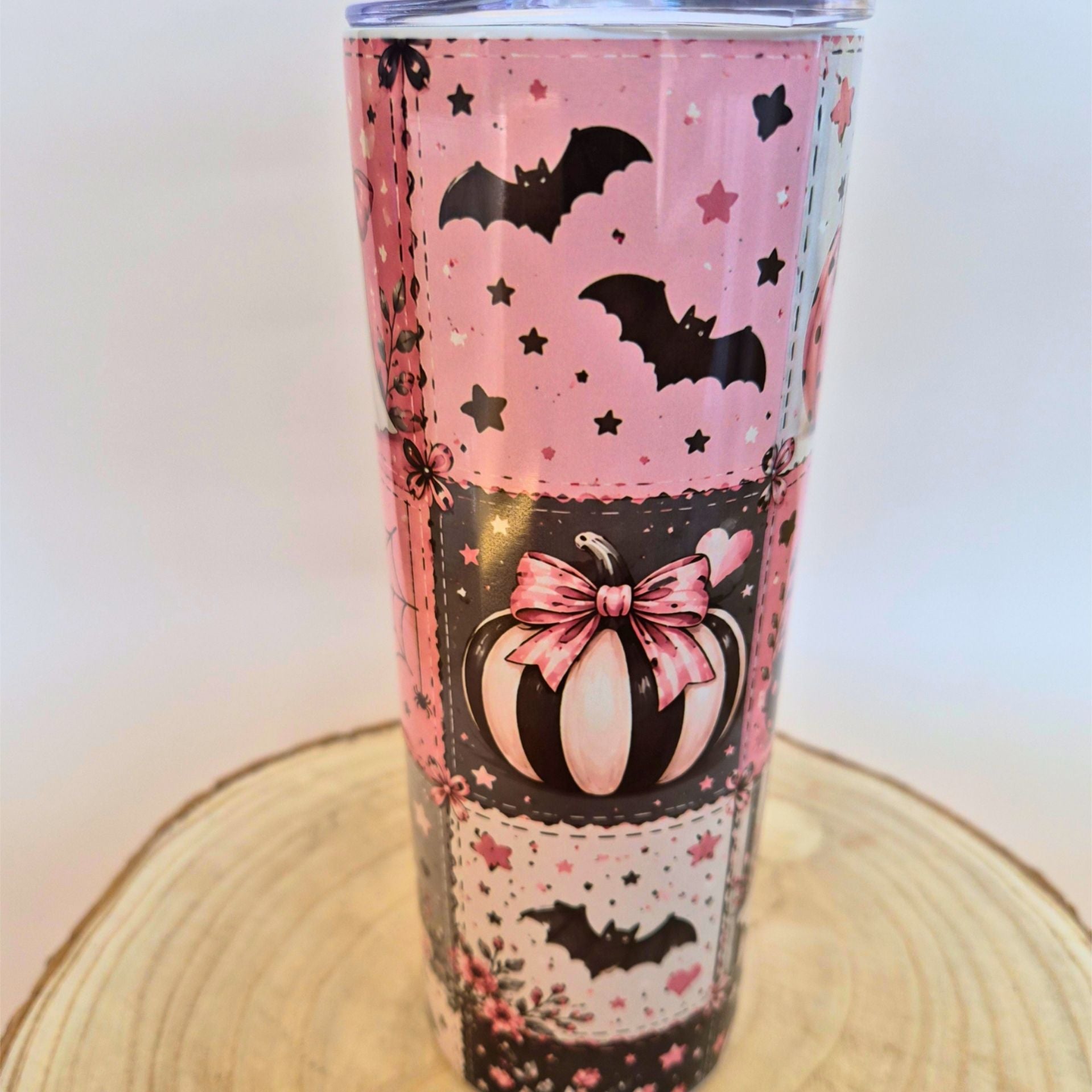A pink and black Halloween-themed tumbler with bats, stars, a pumpkin, and floral lace patterns, shown on a wooden slice coaster against a light background.