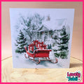 A square greeting card featuring a watercolor painting of a snowy winter scene with a red sleigh carrying red gift boxes, displayed on a wooden surface with a pink border and a 'Lovely Subli' logo in the corner.