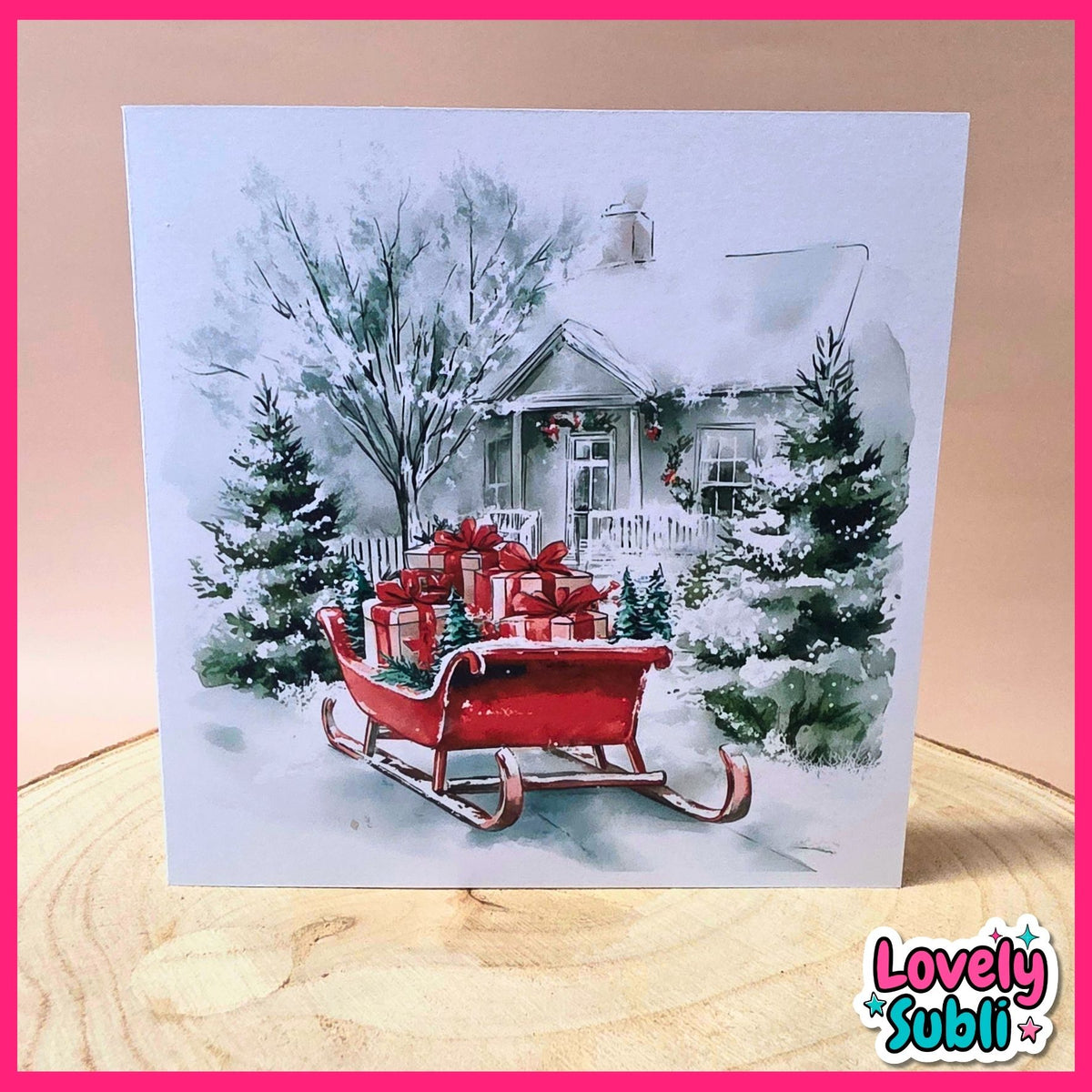A square greeting card featuring a watercolor painting of a snowy winter scene with a red sleigh carrying red gift boxes, displayed on a wooden surface with a pink border and a 'Lovely Subli' logo in the corner.