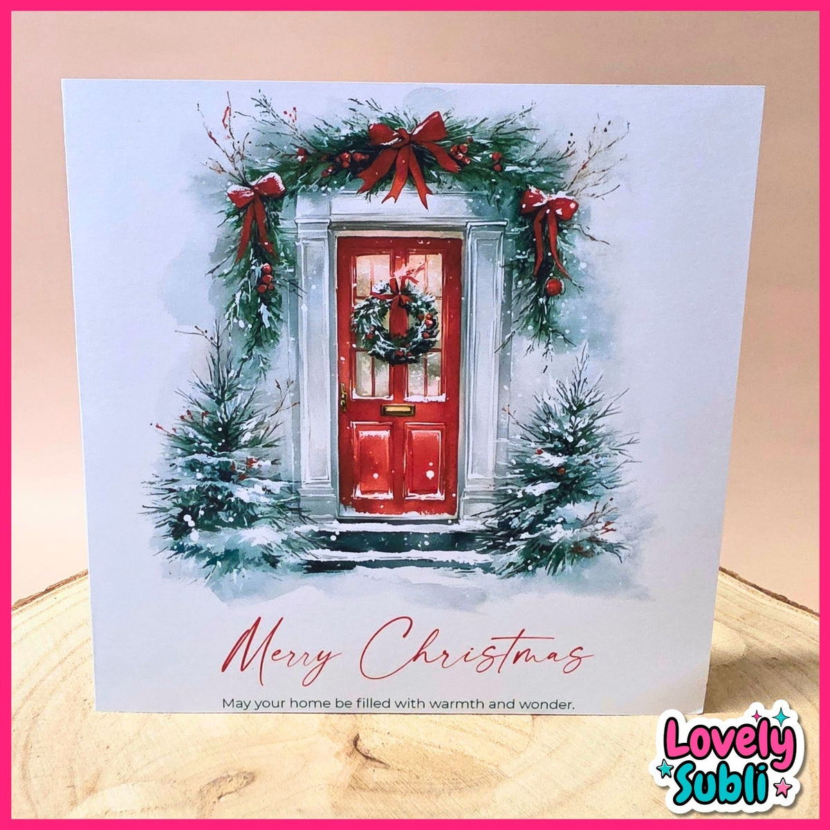 A square Christmas greeting card featuring a painted red front door framed by green garlands, red bows, snow, and small snowy trees, with the text 'Merry Christmas' and a small quote below. The card is photographed standing on a light wood surface against a pale pink background, with a 'Lovely Subli' logo in the bottom-right corner.
