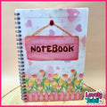 A spiral-bound notebook with a decorative cover featuring a pink hanging sign that says 'NOTEBOOK' and a watercolor-style flower bed with pink, yellow, and orange flowers. The cover has a white background with faint pink hearts.