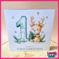 A square baby first Christmas card displayed on a wooden surface. The card features an illustration of a cute baby deer holding a small Christmas tree next to a large number one, with the text 'BABY'S FIRST CHRISTMAS' at the bottom.