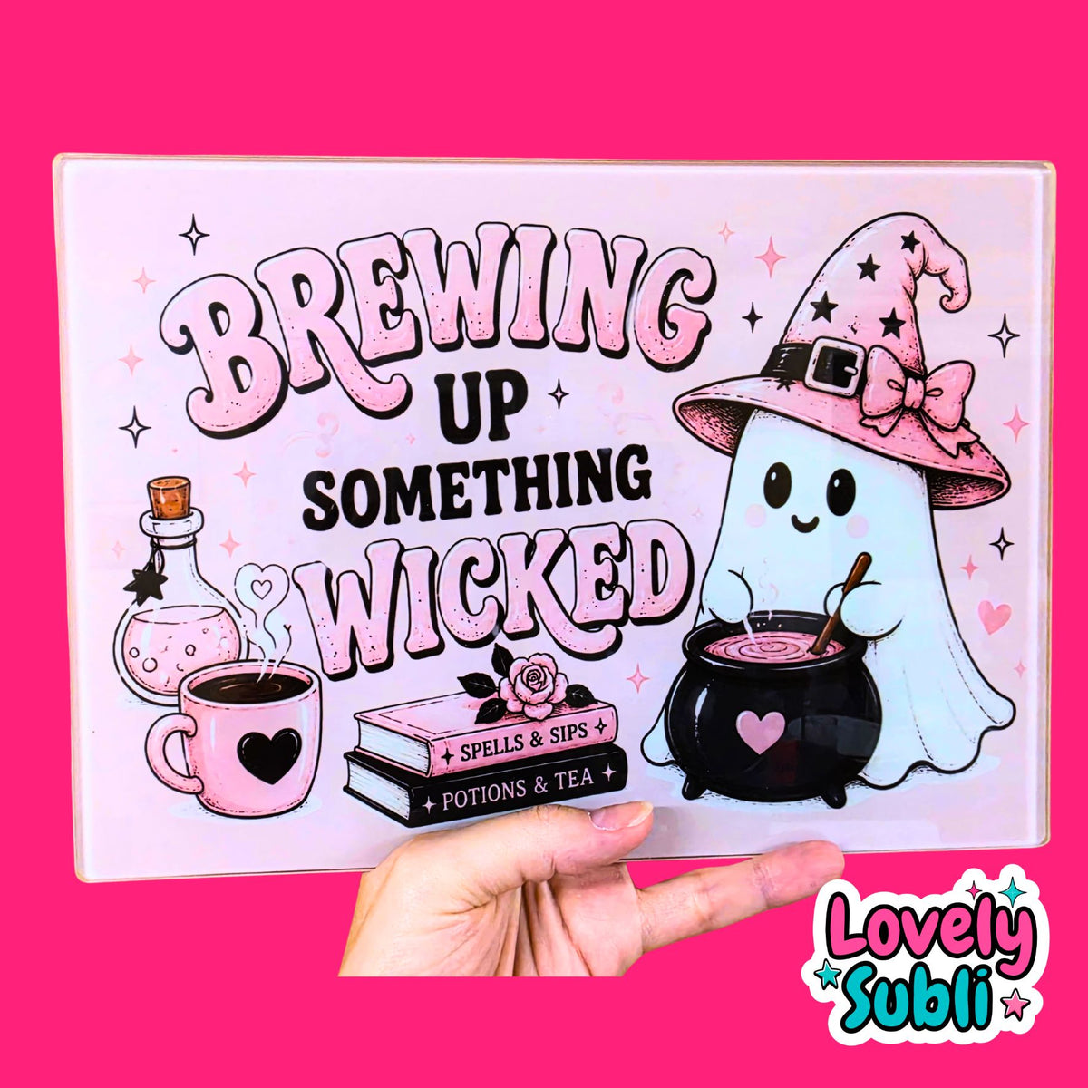 A hand holds a square decorative sign or print featuring a pink and white Halloween-themed design with the text 'BREWING UP SOMETHING WICKED,' a ghost in a witch hat by a cauldron, a potion bottle, a mug, and a stack of books. The background is solid bright pink, and a 'Lovely Subli' logo appears in the bottom right corner.