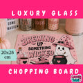 A pink Halloween-themed glass cutting board with a cute ghost and witch illustration, displayed on a countertop with decorative jars and a pink appliance in the background. The image includes promotional text overlays reading 'LUXURY GLASS', '20x28 cm', and 'CHOPPING BOARD'.