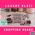 A pink and black Halloween-themed glass cutting board displayed on a white marble countertop with kitchen items in the background. The board features a cartoon ghost in a witch hat, a teapot, a mug, and the text 'CAFFEINE IN MY CUP MAGIC IN MY SOUL CHAOS IN MY HOUSE'.