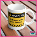 A white ceramic mug with a humorous warning label graphic that reads 'WARNING Not great with other humans' in black and yellow, placed on a brown wooden slatted surface.