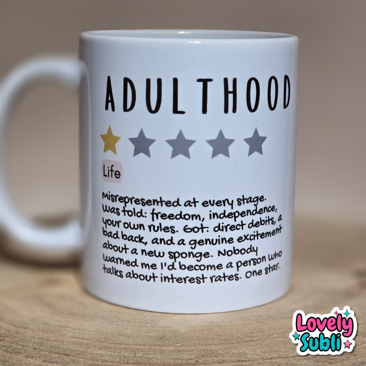A white ceramic mug printed with the word 'ADULTHOOD' in bold black letters, a one-star rating graphic, and a humorous definition of adulthood written in small black text. The mug sits on a wooden surface against a warm beige background.