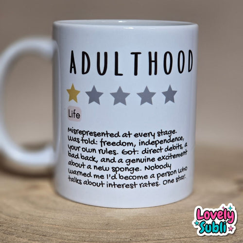 A white ceramic mug printed with the word 'ADULTHOOD' in bold black letters, a one-star rating graphic, and a humorous definition of adulthood written in small black text. The mug sits on a wooden surface against a warm beige background.