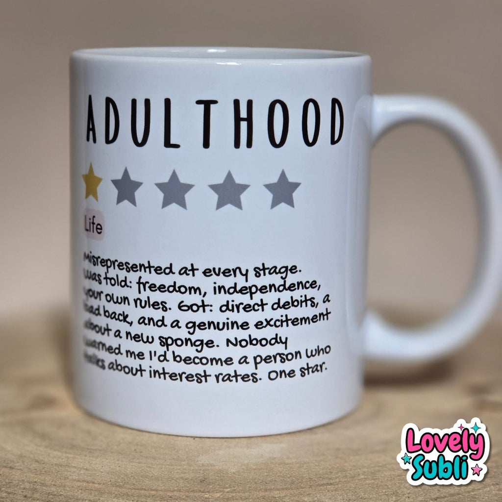 A white ceramic mug printed with the word 'ADULTHOOD' in black, a one-star rating graphic, and a humorous dictionary-style definition. The mug sits on a light wooden surface against a warm beige background.