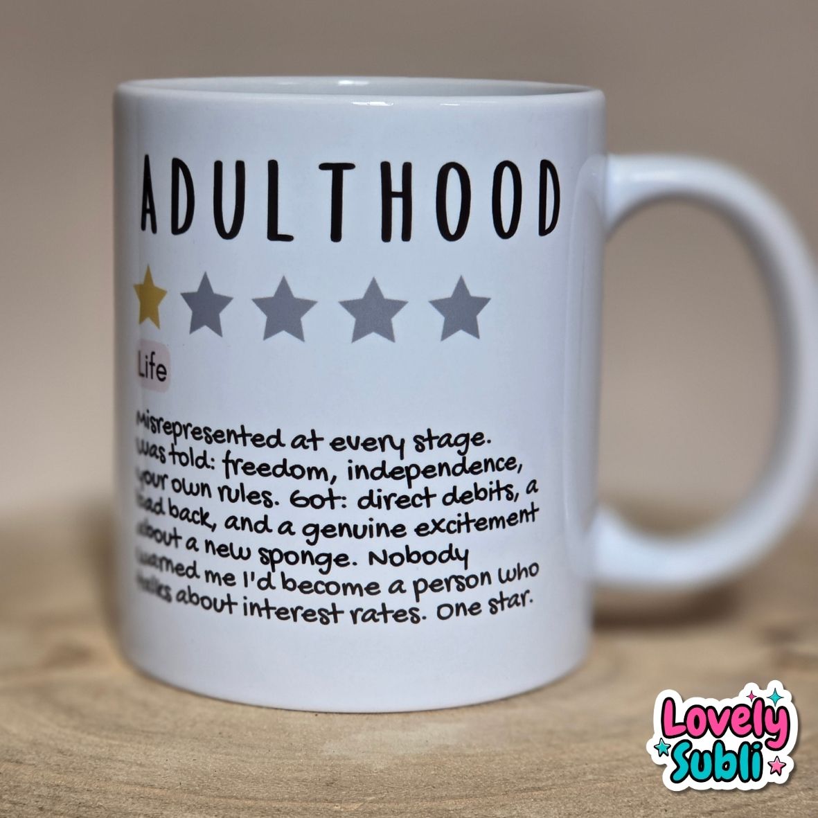 A white ceramic mug printed with the word 'ADULTHOOD' in black, a one-star rating graphic, and a humorous dictionary-style definition. The mug sits on a light wooden surface against a warm beige background.