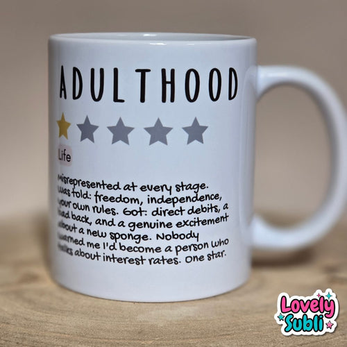 A white ceramic mug printed with the word 'ADULTHOOD' in black, a one-star rating graphic, and a humorous dictionary-style definition. The mug sits on a light wooden surface against a warm beige background.