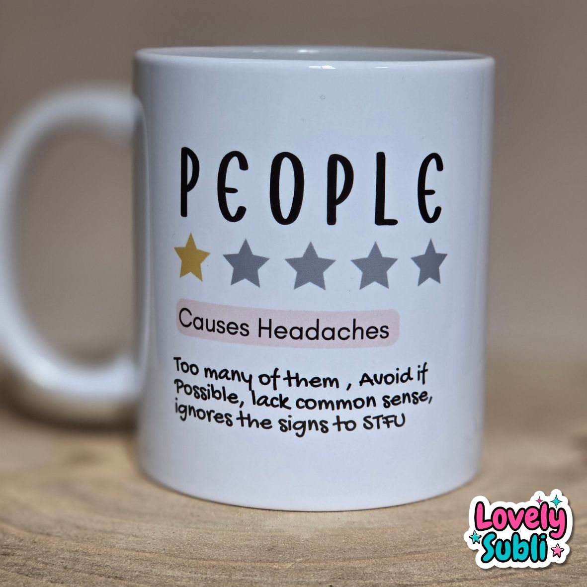 A white ceramic mug printed with the word 'PEOPLE' in black, five stars (one gold, four gray), and a humorous definition of people causing headaches, placed on a wooden surface with a blurred beige background.