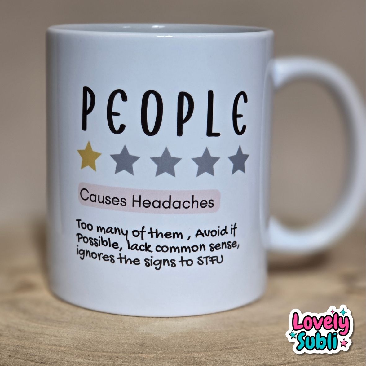 A white ceramic mug printed with the word 'PEOPLE' in black, five stars (one gold, four gray), the phrase 'Causes Headaches' in black and pink, and a humorous definition below. The mug is photographed on a wooden surface with a warm beige background.
