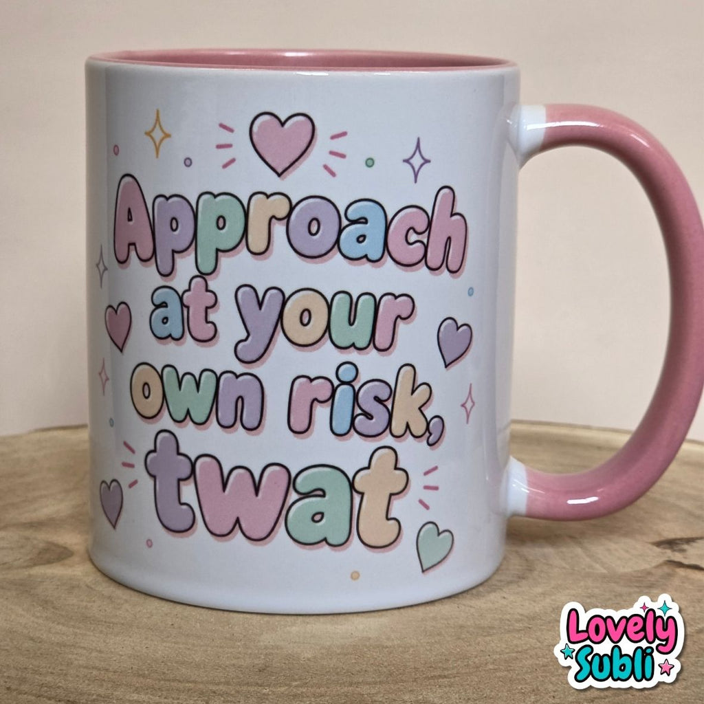 A white ceramic mug with a pink handle and interior, featuring the text 'Approach at your own risk, twat' in pastel multicolor letters with heart and star accents. The mug is photographed on a wooden surface against a plain beige background, with a 'Lovely Subli' watermark in the bottom right corner.