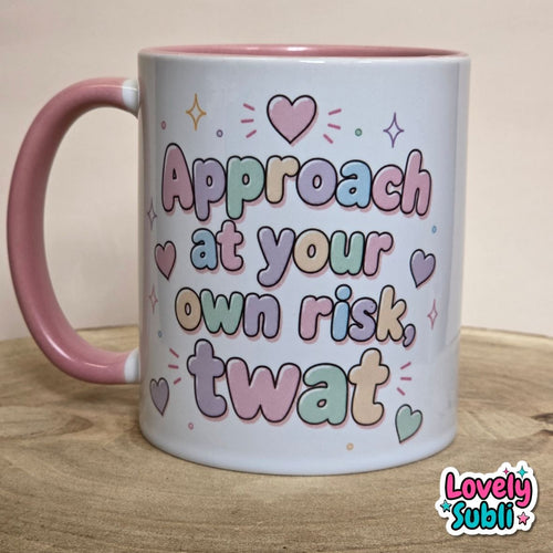 A white ceramic mug with a pink handle and interior, featuring the text 'Approach at your own risk, twat' in pastel multicolor lettering with heart and star accents. The mug sits on a wooden surface against a neutral beige background, with a 'Lovely Subli' watermark in the bottom right corner.
