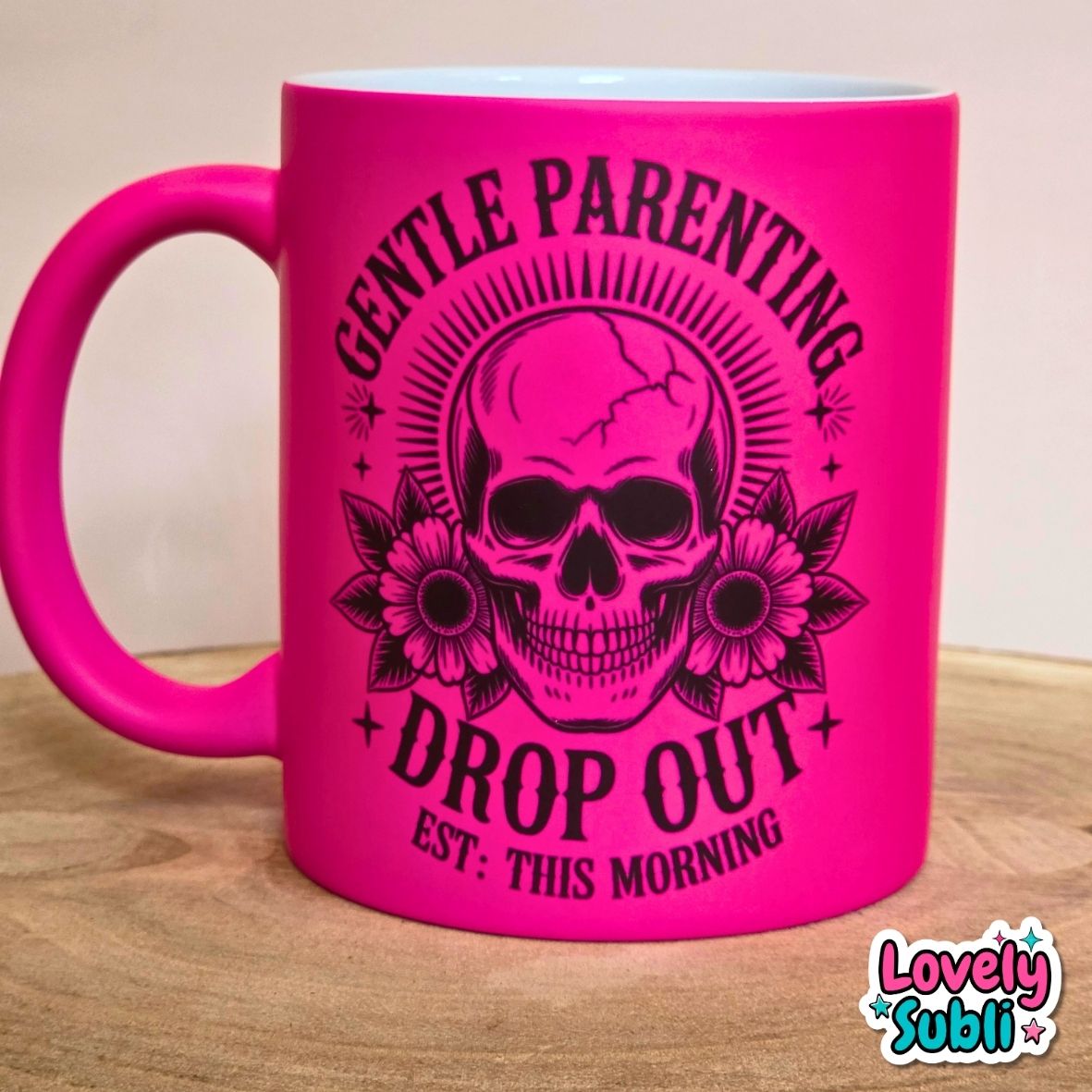 A bright hot pink ceramic mug with a black graphic of a skull, flowers, and the text 'GENTLE PARENTING DROP OUT EST: THIS MORNING' on a wooden surface.
