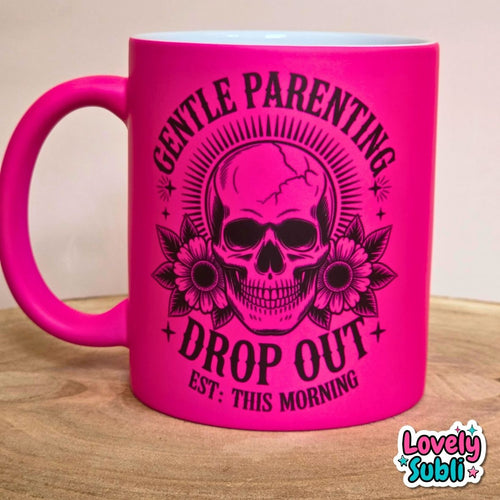 A bright hot pink ceramic mug with a black graphic of a skull, flowers, and the text 'GENTLE PARENTING DROP OUT EST: THIS MORNING' on a wooden surface.