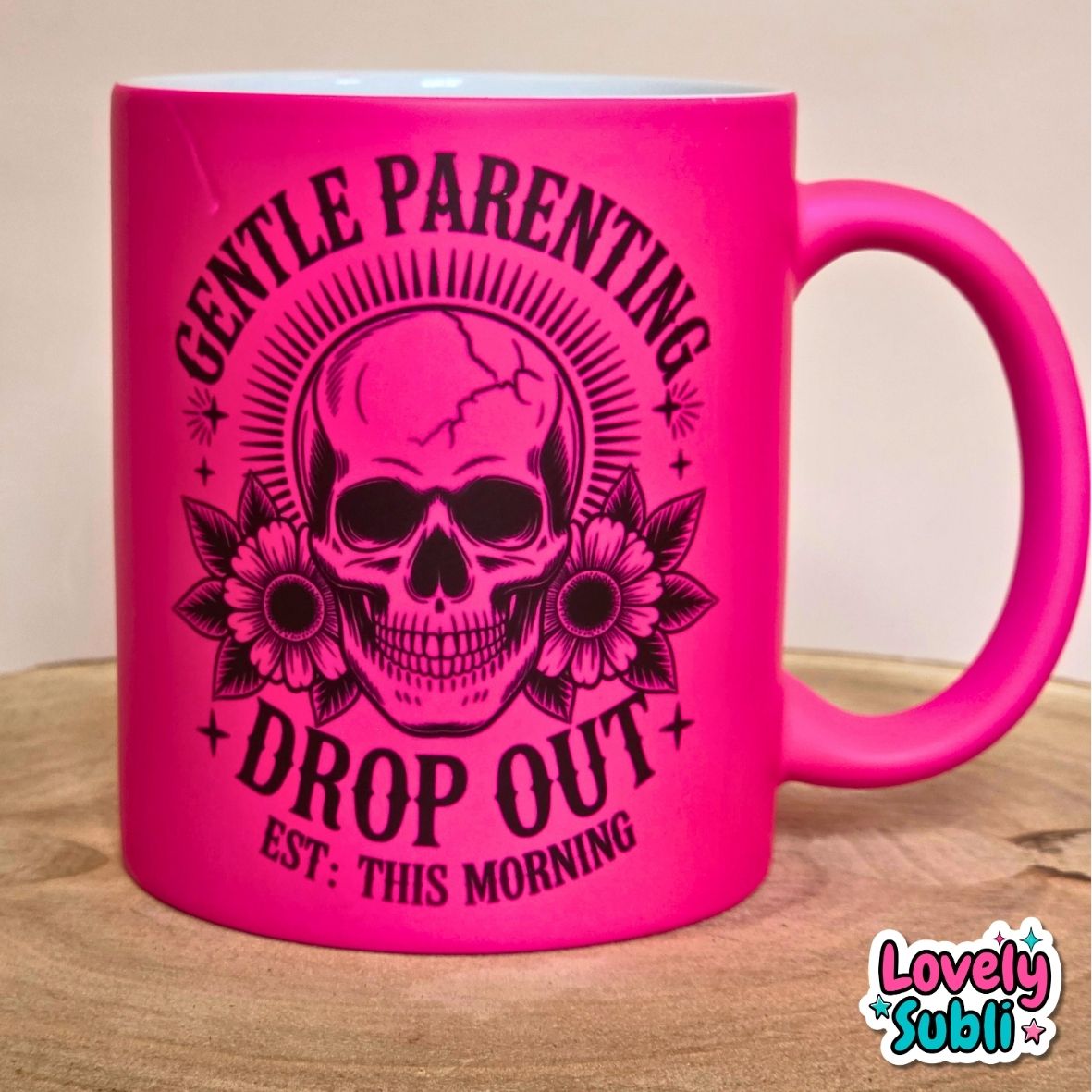 A bright hot pink ceramic mug with a black graphic of a skull wearing ear cuffs, surrounded by flowers and the text 'GENTLE PARENTING DROP OUT EST. THIS MORNING'.
