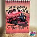 A spiral-bound notebook with a pink and red cover featuring an illustration of a black steam train and the text 'I'M NOT ALWAYS A TRAIN WRECK Just kidding TOOT TOOT'. The notebook is standing on a wooden surface against a neutral beige background.