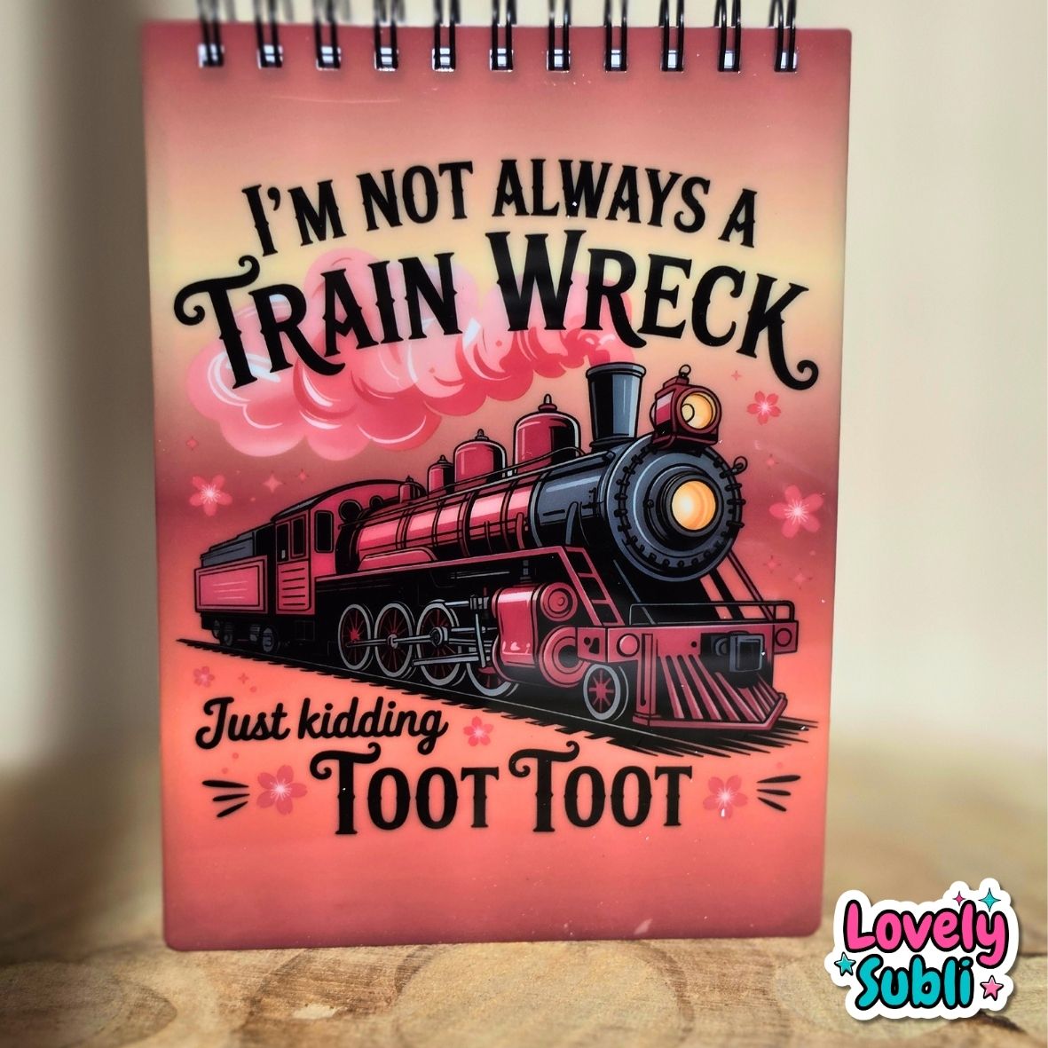 A spiral-bound notebook with a pink and red cover featuring an illustration of a black steam train and the text 'I'M NOT ALWAYS A TRAIN WRECK Just kidding TOOT TOOT'. The notebook is standing on a wooden surface against a neutral beige background.