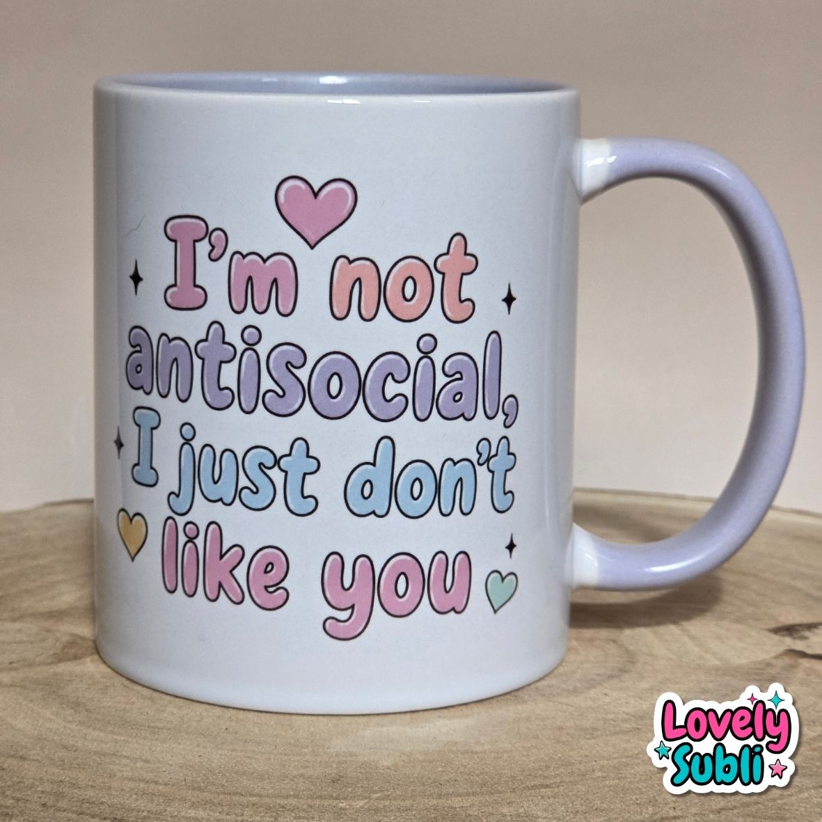 A white ceramic mug with a pastel message that reads 'I'm not antisocial, I just don't like you' in pink, purple, and blue lettering, accompanied by small heart and star accents. The mug is placed on a wooden surface against a neutral beige background, with a 'Lovely Subli' brand logo in the bottom right corner.