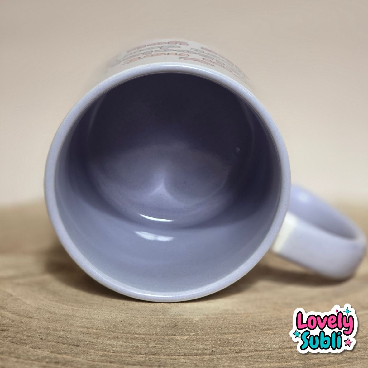 A lilac ceramic mug shown from a top-down angle, lying on its side on a wooden surface with a beige background. The inside of the mug is a dusty lavender-blue, and the handle is on the right.